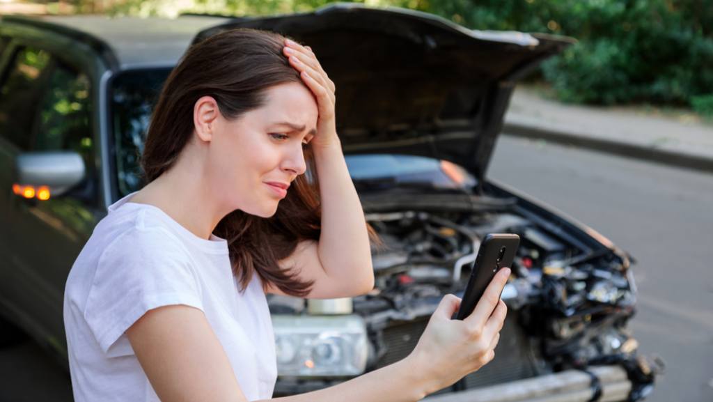 Distressed woman clutching head and checking black cellphone after car crash dark vehicle in background daytime
