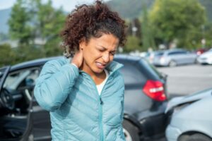 Woman standing rubbing neck and grimacing after car crash black car in background rear end collision with silver car
